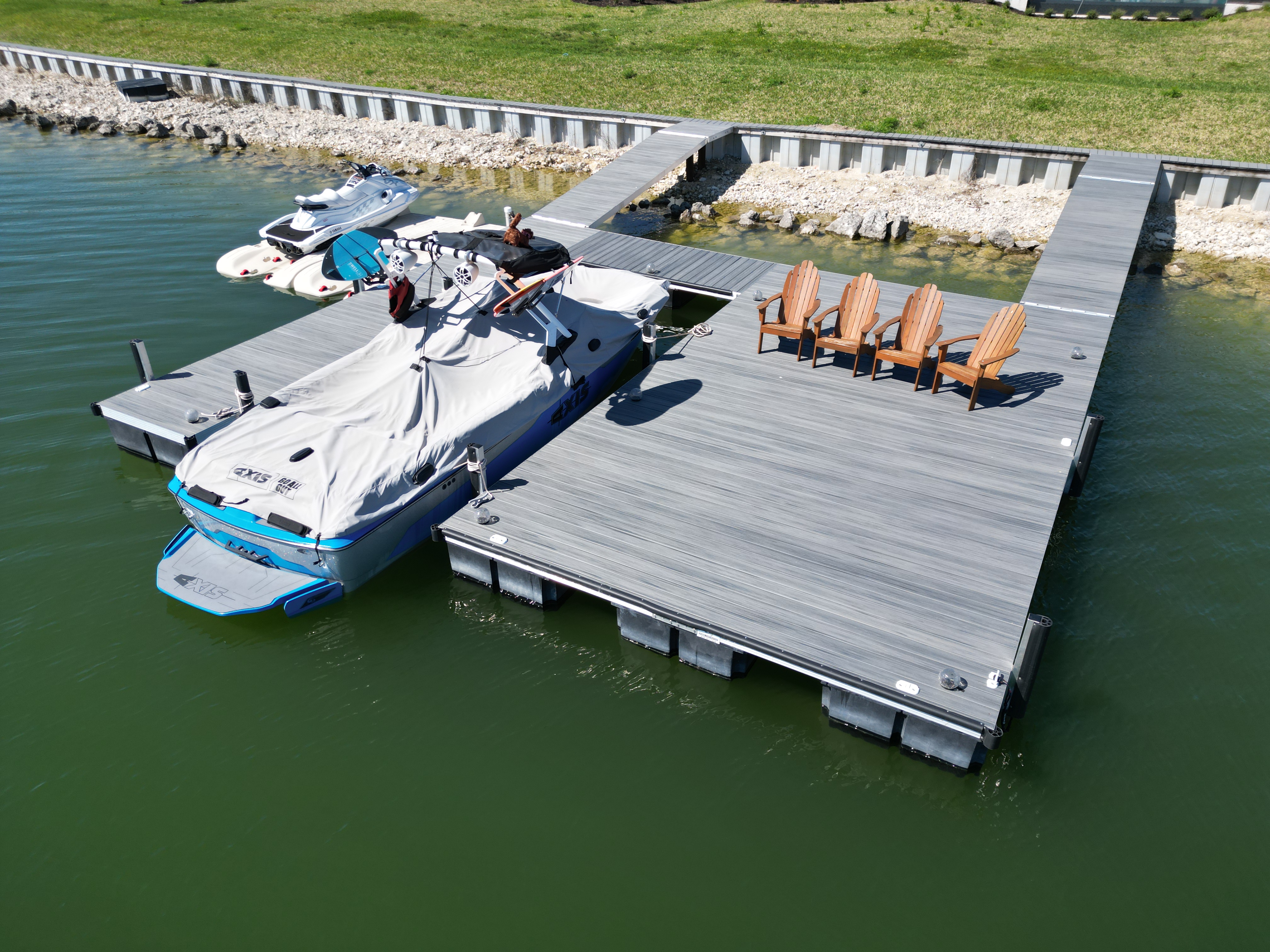 Aerial view of a waterfront dock with a covered boat and jet skis. The gray floating dock extends over green water, with wooden chairs lined up on the right side. A boat with a white cover is tied alongside the dock, and a rocky shoreline with grass is visible in the background.