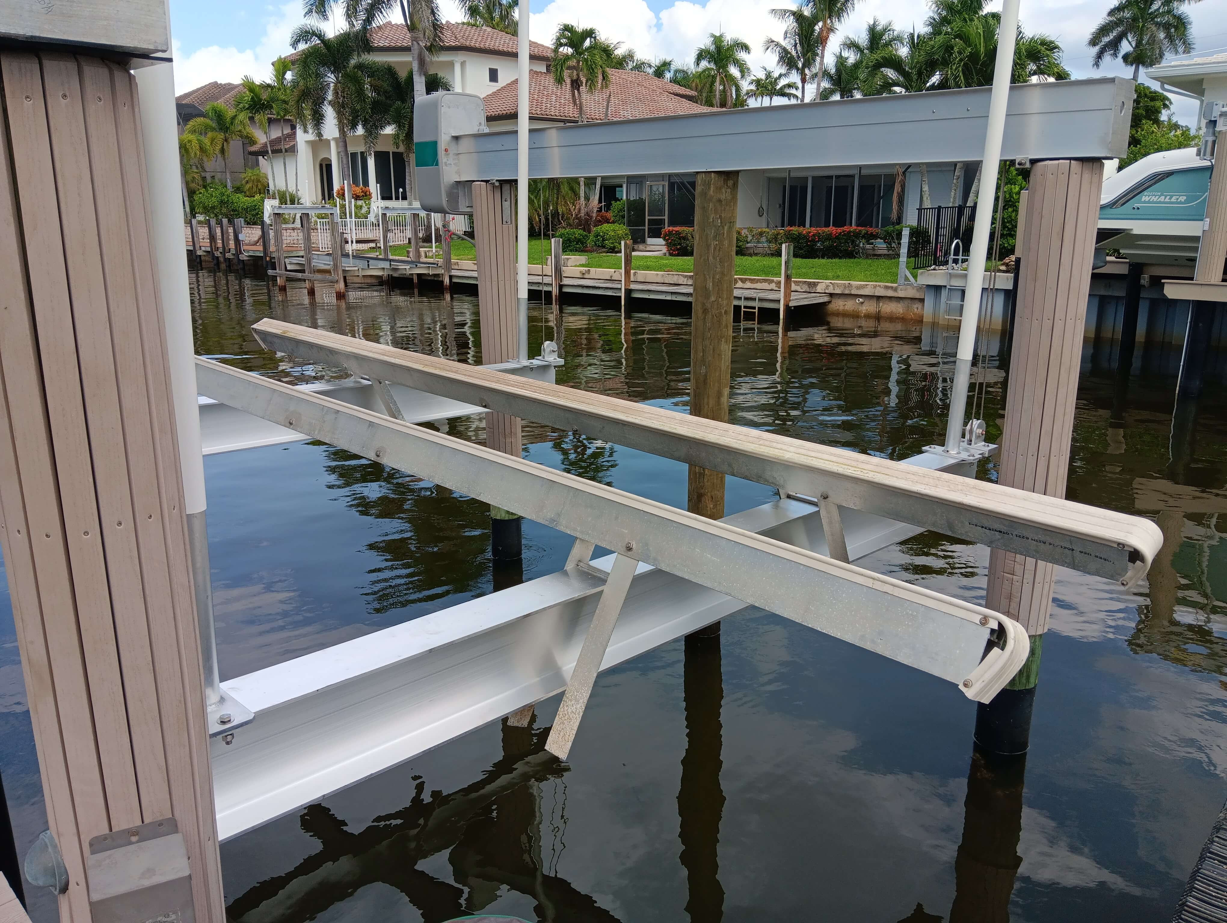 Boat lift at a waterfront dock with white metal frame, supports, and calm water surrounded by homes and palm trees.